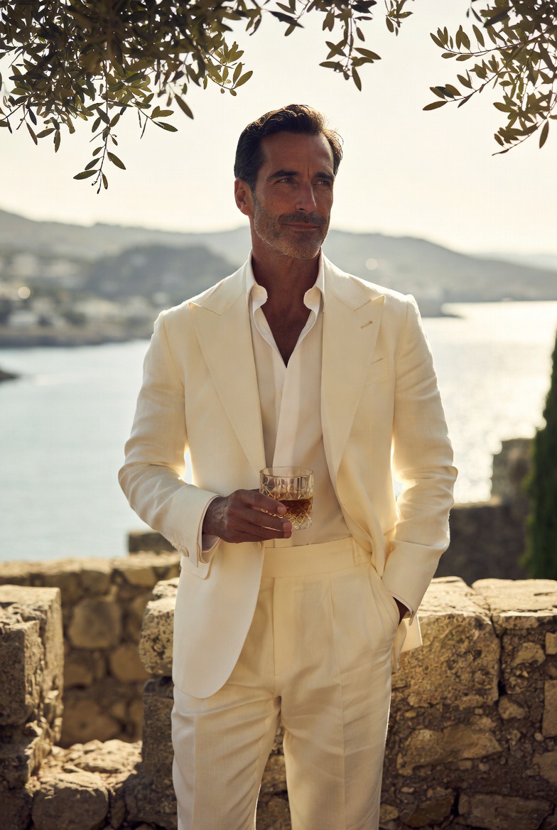 Hombre con traje crema sosteniendo una copa de whisky junto al mar Mediterráneo